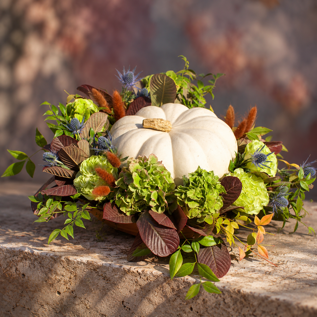 Green hydrangeas, ruffled lime-green blooms, and blue thistle ring a white pumpkin with smokebush. A fall centerpiece by Empty Vase in Los Angeles.