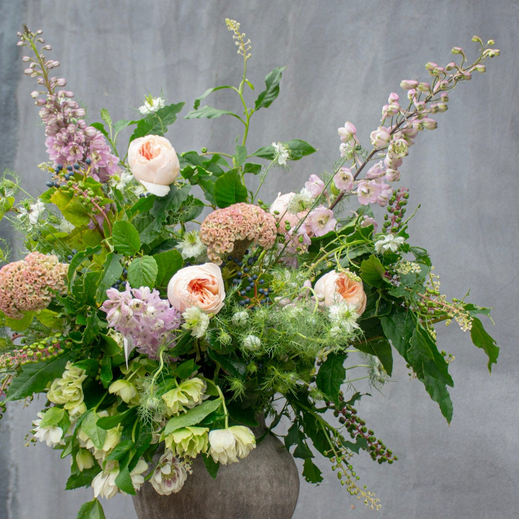 A lush wildflower arrangement in a rustic gray ceramic vase, displayed on a wooden stool against a textured gray wall.