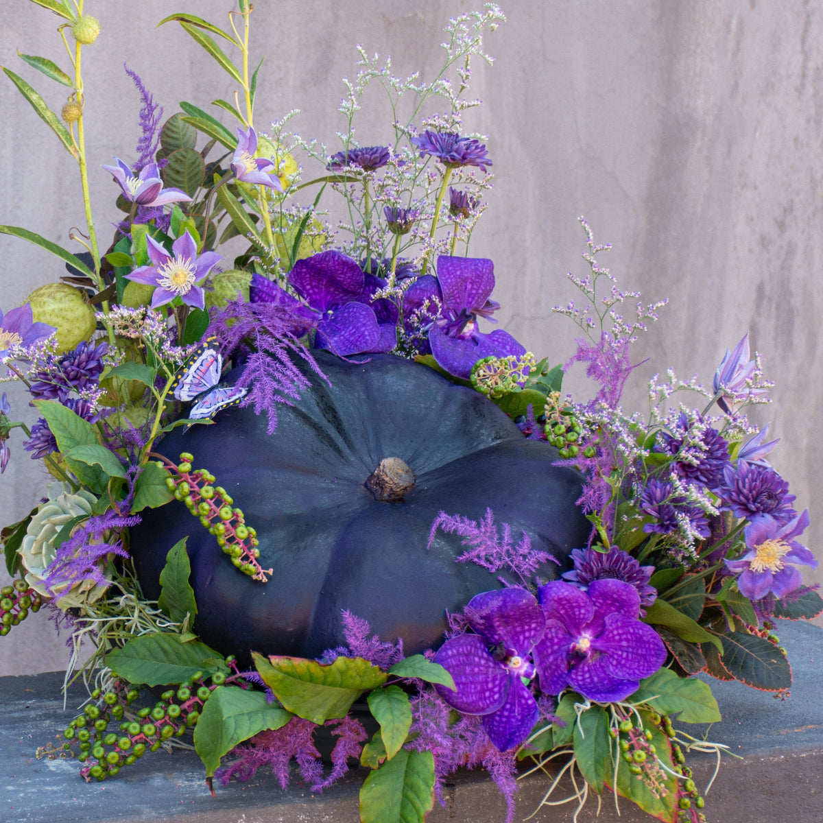 Amethyst Garden arrangement of purple Vanda orchids, clematis, and astilbe around a plum pumpkin in a black-painted terracotta vessel